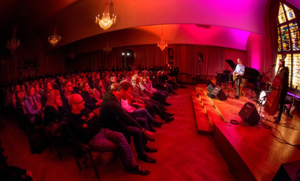 Publikum schaut einen Musiker auf der Bühne in einem rot beleuchteten Raum an.